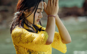身穿黃色蕾絲上衣的年輕女性在水邊將雙手交握在臉龐附近