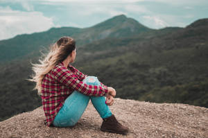 穿著格仔襯衫和牛仔褲的女性坐在山頂上俯瞰森林覆蓋的山丘。