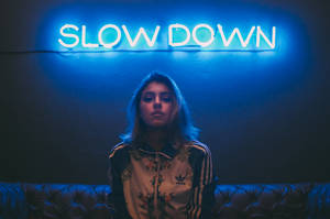 穿著 adidas 外套的女性坐在藍色霓虹燈牌下，燈牌寫著 slow down。