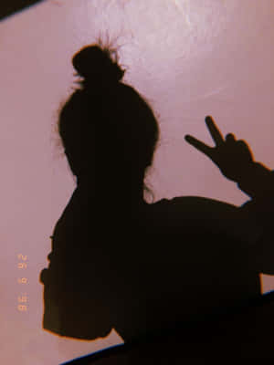 一個束著髮髻的人影，在粉紅色牆前做出和平手勢。