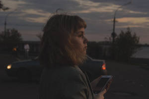 女性在黃昏的街道上手持電話，身後有汽車駛過。 — 深色iPhone壁紙