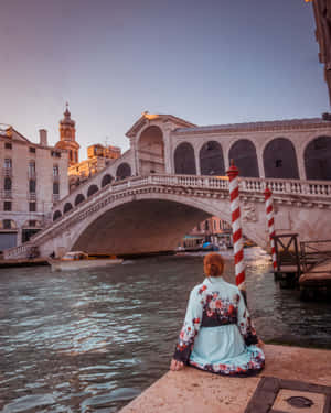 身穿花卉和服的人坐在威尼斯運河邊，觀看夕陽下的里亞爾托橋。