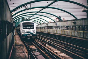 一列火車在都市環境中駛近綠色金屬天篷下的高架軌道。