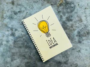 米色筆記本，標有「IDEA NOTEBOOK」，在灰色混凝土上有黃色燈泡圖案。 — 勵志桌面 壁紙