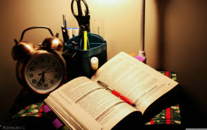 書桌上有打開的書、紅色筆、鬧鐘和散發溫暖光線的檯燈。