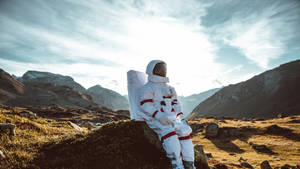 身穿白色太空衣的太空人坐在岩石地形上，背景是日落時分的山脈。 — 太空人 壁紙