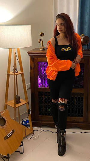 穿著橙色外套和黑色服裝的女性站在有結他和霓虹櫥櫃的房間裡。