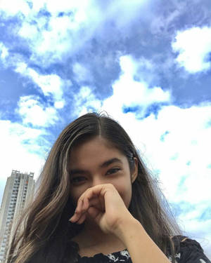 年輕女性在戶外多雲的天空下，背景有建築物，頑皮地捂住嘴巴。