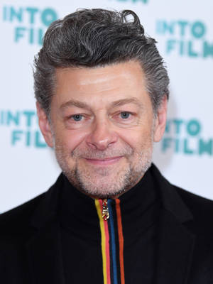 演員安迪·瑟克斯（Andy Serkis）在活動中穿著帶有彩色拉鏈的黑色外套微笑。 — 荷里活 GIF 壁紙