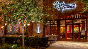 Roy's Hawaiian Fusion Cuisine 餐廳夜晚入口，帶有亮燈招牌和開花的樹。