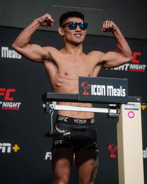 UFC拳手在稱重活動中站在體重秤上展示肌肉，戴著太陽眼鏡和品牌短褲。