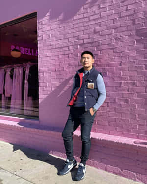 穿著街頭服飾的男子靠在粉紅磚牆上，鄰近服裝店。