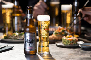 朝日啤酒瓶和玻璃杯，配以壽司碟和筷子，在餐廳環境中。 — 日本 壁紙