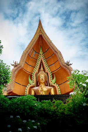 金色佛像在裝飾寺廟屋頂下，被綠色植物和雲朵環繞。