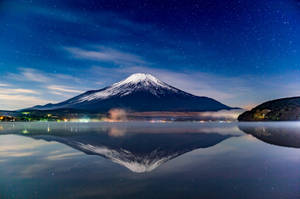 富士山在星空下平靜的湖面上倒映，遠方燈光點點。