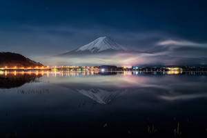 富士山在星空下的平靜湖面倒映，遠處有城市燈光。