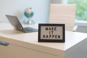 一塊寫有「make it happen」的字母板放在白色桌上，旁邊有手提電腦和地球儀。 — 勵志桌面 壁紙