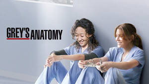 兩位穿著藍色手術服的女人微笑著坐在牆前，牆上有「Grey's Anatomy」文字。