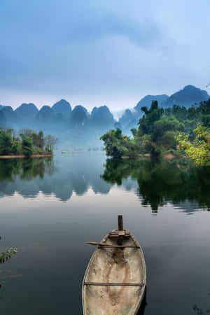 一艘小船停泊在平靜的河面上，四周環繞著霧氣繚繞的山脈和茂密的綠植。 — Huawei MatePad 12 X (2025) 壁紙