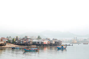 漁船漂浮在高腳屋附近，遠處山巒環繞的霧氣瀰漫海邊村莊。 — 河景 壁紙