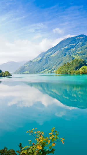 平靜的綠松石色湖泊反射山脈和天空，前景有秋葉 — 安卓 壁紙