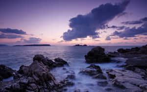 紫色天空同平靜嘅海浪喺日落時拍打黑色岩石。