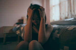 年輕女性坐在床上雙手抱頭神情痛苦在光線昏暗的房間 — 抑鬱 GIF 壁紙