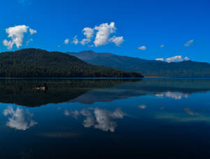 平靜的山湖反射出藍天白雲，背景是森林覆蓋的山丘。 — 湖景 壁紙