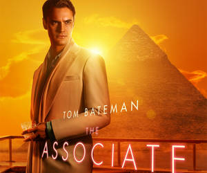 Tom Bateman 身穿米色西裝，在夕陽下的金字塔前拿著飲品。