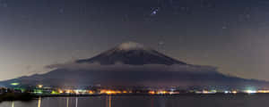 富士山在星空下，城市燈光在夜間倒映在湖面上 — 雙螢幕 壁紙