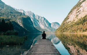 一個人獨自坐在木碼頭上，俯瞰平靜的湖泊，背景有山脈。 — 旅行 壁紙