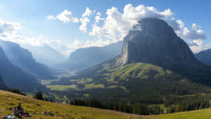 兩個人在長滿草的山丘上休息，俯瞰山谷和一座大山峰，天空晴朗。