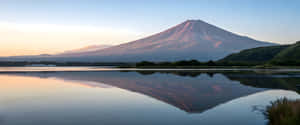 富士山在日出時倒映在平靜的湖泊中，背景是綠色山丘