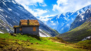 一座小木屋坐落在山谷中，周圍是白雪皚皚的山峰和綠草。 — 山景 壁紙