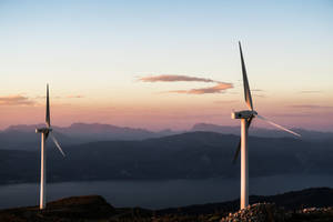 夕陽下，兩座風力發電機位於俯瞰山巒和海洋的山丘上。