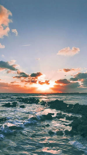 海洋上的夕陽，海浪拍打岩石，雲朵閃耀橙色光芒 — 沙灘 壁紙