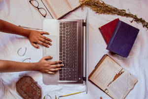 Hands typing on a laptop surrounded by books and dried flowers on a bed.