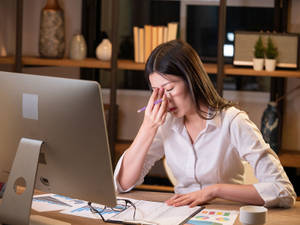 一位女性在辦公桌前盯著電腦螢幕和圖表後揉眼睛。