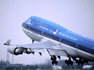 藍色KLM波音747飛機降落，起落架清晰可見，背景為城市。