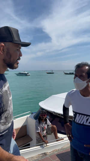 三個男人喺熱帶碼頭嘅船上，周圍有藍綠色海水同遠處嘅船。 — 運動 壁紙