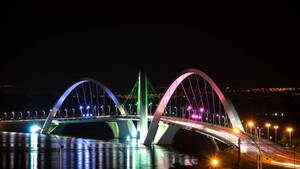 夜間一座明亮照明的現代橋樑橫跨水面，背景有車流痕跡和城市燈光。 — 通往泰拉比提亞之橋 壁紙