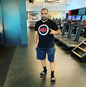 男子在健身室指著AKA Wrestling T恤，附近有跑步機。