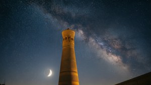一座歷史悠久的宣禮塔在夜晚被照亮，星空下有一彎新月和銀河。