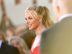 Britney Spears 身穿紅色連衣裙在活動中微笑，背景人群模糊。