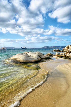 碧綠的海浪沖刷著沙灘，沙灘上有大岩石和船隻，天空多雲而湛藍。 — 海浪 壁紙