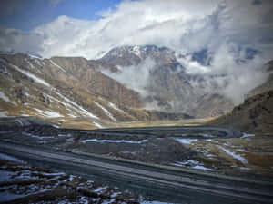 蜿蜒的山路，帶有雪斑和雲朵，背景是崎嶇的山峰。