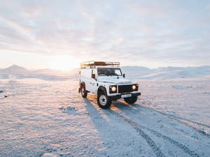 白色 Land Rover Defender 在日出時分駛過雪覆蓋的沙漠，背景有山。