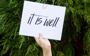 手舉著寫有「it is well」的標牌，背景為綠色植物。