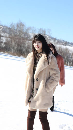 穿米色外套的女子在雪地中微笑，背景有光禿樹木和山丘。