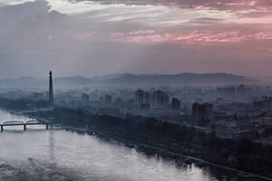 一條河流在黎明時分蜿蜒穿過霧氣瀰漫的城市，背景中有一座高大的紀念碑和山脈。